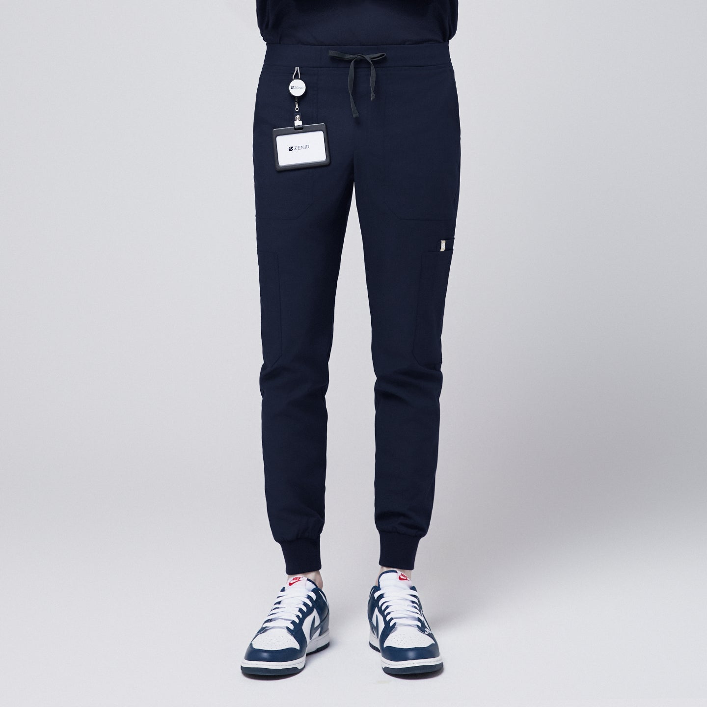 Image of Front view of navy jogger scrub pants with 8 pockets, drawstring waistband, cuffed ankles, and a name badge, paired with navy sneakers,Eco Navy